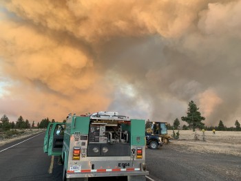 A photo of the back of a wildland fire engine stopped on a road. Smoke in the background.