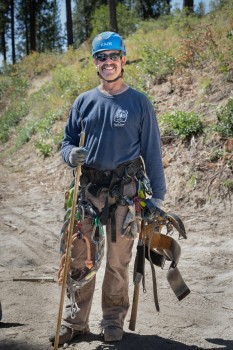 A man wears a helmet, harness, and is holding tree climbing gear. 