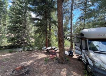 Twp campers sit by an RV in a forest near a river.
