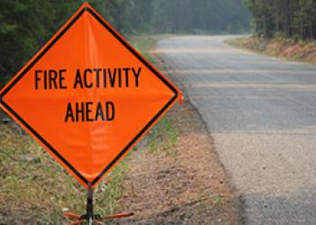 A photo of a Fire Activity Ahead sign next to a paved road in a forest.