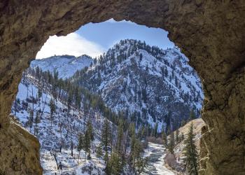 A photo taken from inside a cave, looking out towards a creek and snowy mountains.