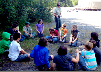 A group of children sit on the ground in a circle as an instructor walks and talks.