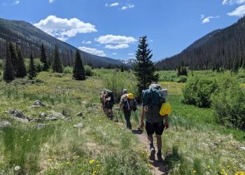 Several volunteers wearing backpacks and carrying helmet and tools walk up a mountain trail. 