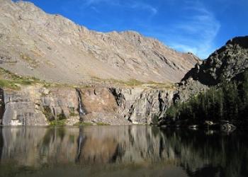 A small lake sits below steep slopes and cliffed shores of a mostly vegetation free mountain. willow lake _Sangre de Cristos.
