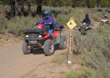 Three people ride on small four-wheel drive vehicles on a dirt trail. 
