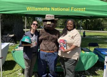 Two individuals with Smokey Bear in front of a Willamette National Forest booth holding Smokey Bear materials.