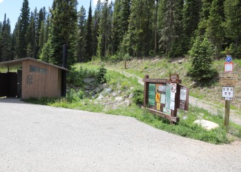 A photo of a Trailhead sign, restroom, and parking lot pavement. A dirt trail leads from the parking lot behind the sign and restroom. Pine trees line the hillside behind the trail.