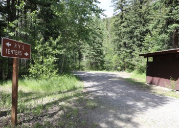 A photo of a brown sign that has an arrow pointing to the left that states, "RV's" and an arrow pointing to the right that states, "Tenters". To the right of the sign is a dirt road, a bathroom facility and a small dirt parking lot. Pine trees surround the area.