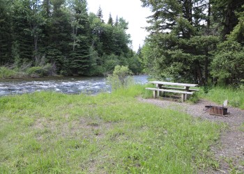 A photo of a picnic table and metal campfire ring near a grassy area that overlooks a river and pine trees line the river.