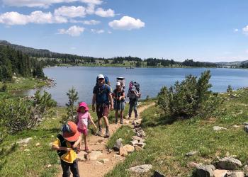 A family of five hiking along a dirt trail. Two small children in front of the group. A large lake is behind them.