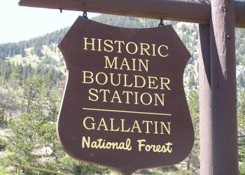 A photo of a wooden sign hanging from a wood post. The sign states, Historic Main Boulder Station Gallatin National Forest