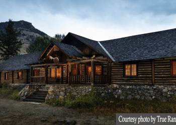 A photo of a wide cabin with about seven windows and a porch, light is shining from the windows.