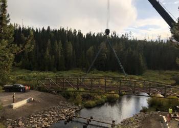A photo of a crane placing a bridge across water into place in the forest.