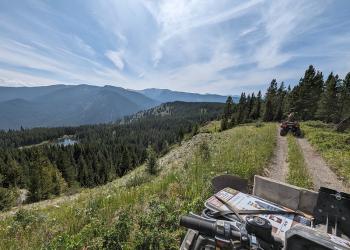 A photo of the front of an OHV on a dirt trail with scenic mountain views. Another OHV rider is ahead on the trail.