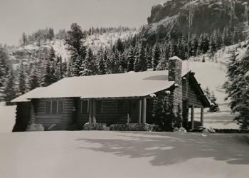 A black and white photo of a cabin in the forest, snow covers the ground and roof of the cabin.