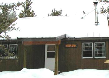 Wooden cabin with metal chimney.  Snow on the ground and icicles dangling off a snow packed roof. .   