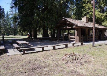 A photo of a rustic kitchen in a timber beam shelter structure with an adjacent paved patio area with picnic tables.