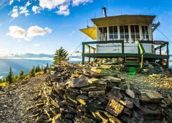 A fire lookout cab with its shutters raised, sits low to the ground on a stony mountain peak with wide vistas behind.