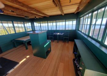 Green-painted furniture and a light colored wooden floor contrast with the walls made of windows in this interior photo of a historic fire lookout.
