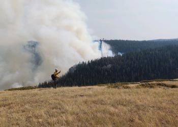 An image of a firefighter taking a weather reading during a fire with smoke and trees behind him