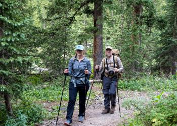 Two hikers pose for the camera as they walk down a forest trail with walking sticks