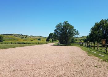 Gravel parking lot surrounded by green grass.  On the right side there are parking stops, a wooden rail fence, gate, and information board inside the campground.