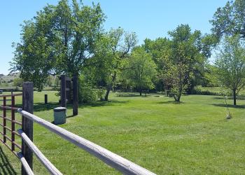 A wooden fence with a red metal gate to the left looking into the campground with green mowed grass and multiple trees scattered through the area.
