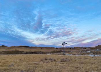 Image is a landscape view of Indian Creek Recommended for Wilderness Area. It shows a windmill with rolling hills in the background. The ground is covered with grass that is yellow in color. The sky is blue with some clouds. 