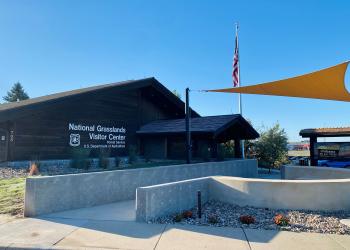 Exterior view of the National Grasslands Visitor Center showing a brown building with a semi-circle amphitheater made from concrete. 