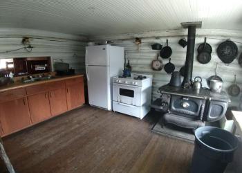 Kitchen with cabinets, propane refrigerator and stove and a antique wood fired stove oven with cast iron pans hanging on a white painted log wall.