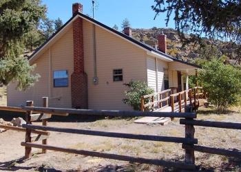 End view of a beige cabin with an old brick fireplace set in front of a buck and pole fence under a pair of trees.  ADA ramp visible to front door with hills and a blue sky behind the cabin.