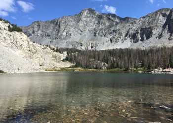 Looking across a calm small lake to the trees on the opposite shore with steep mountains rising up behind them. 