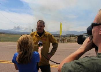 A man in a yellow fire shirt stands in front of a woman in a blue shirt holding a microphone while a cameraman films the pair with the fire smoke in the distance.