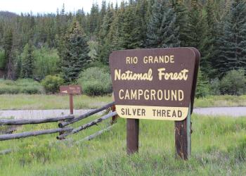 The brown and tan Silver Thread campground sign stands in a grassy area with a pole fence behind it. 