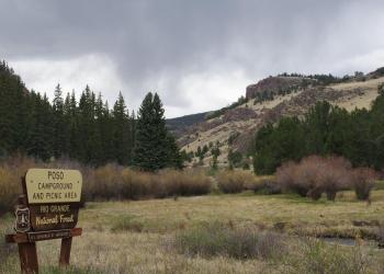 The brown and tan Poso campground sign on the left with  small meadow in the foreground and shrubs, trees and cliffs behind. 