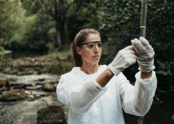 A woman wearing a white protective suit holds up a vile . A stream and Forest are in teh background. 