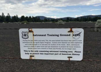 A rusted white metal sign reading Astronaut Training Ground with descriptive test hangs from a barbed wire fence with a cinder field, pine trees and mountains in the background