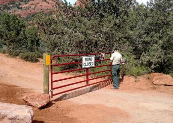A uniformed Forest Service employee stands in front of a red gate with a sign reading ROAD CLOSED positioned over a dirt road with green trees in the background