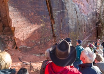 A person with a brown cowboy hat stands among a larger group of people with a shaft of light cutting across a red rock wall filled with petroglyphs
