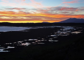 A orange and yellow sunset is visible above a mountain range and a shallow pond