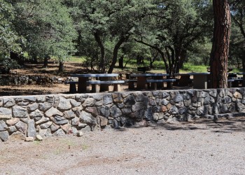 A rock wall separates a gravel parking area from shaded picnic tables