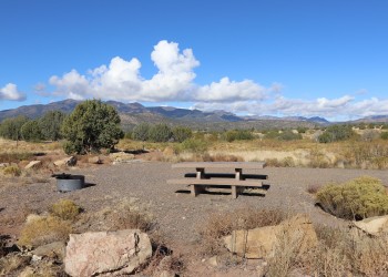 Photo of a graveled campsite with picnic table and fire ring with mountains in the background under a blue sky