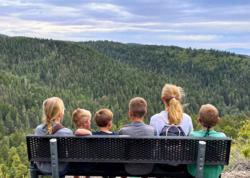 six people on a bench looking out towards mountain and trees