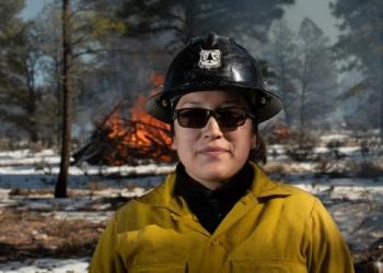 A woman wearing a black plastic helmet, sunglasses, and a yellow fire shirt standing in a snowy forest faces the viewer while a pile of woody debris burns behind her.