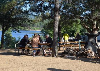 People at picnic tables in the shade with lake in background.