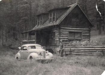 A person stands in front of a historic recreation residence cabin in the 1950s
