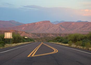 This photo shows the roadway while a pink sunset-colored mountains are spotted in the background below a multicolored sky