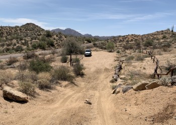 Small trail entrance with boulder perimeter, jeep parked at the entrance, and mountain background.