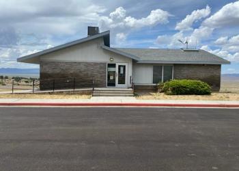 Front of the Austin-Tonopah Ranger District's Austin Office, which is a single-story building with brick and white paneling, door with a ramp, large window, and shrubbery. The sky is partly cloudy, and the ground is paved with asphalt.