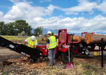 Two workers in hard hats and yellow safety vests feed timber through a firewood processing machine. 